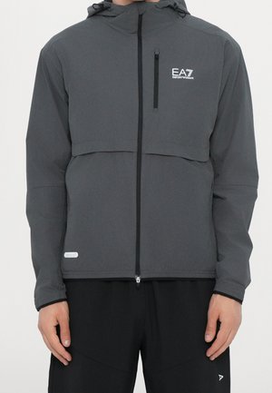 Grå hættejakke med lynlås, brystlomme og EA7 Emporio Armani-logo, båret af en person med hænderne afslappet ned langs siderne.
