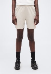 Άτομο που φοράει μπεζ σορτς, μαύρες κάλτσες μέχρι τον αστράγαλο και μαύρα αθλητικά παπούτσια, στέκεται μπροστά σε ένα απλό λευκό φόντο.