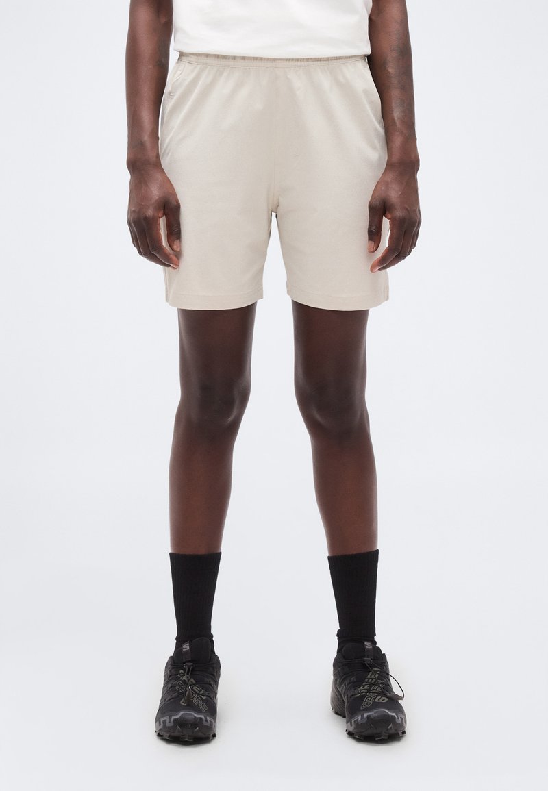 Άτομο που φοράει μπεζ σορτς, μαύρες κάλτσες μέχρι τον αστράγαλο και μαύρα αθλητικά παπούτσια, στέκεται μπροστά σε ένα απλό λευκό φόντο.