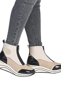 Bottes chevilles beiges et noires au design ressemblant à des chaussettes, avec un bout en vernis et des accents en daim texturé, dotées d'une fermeture éclair sur le côté.
