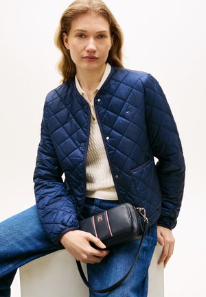 Moteris dėvi tamsiai mėlyną siūlų striukę per kakavinį megztinį ir mėlynas džinsus, laiko juodą per petį nešiojamą rankinę su raudonais ir baltais dryžiais.