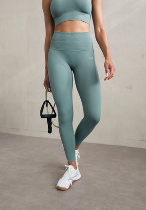 Ensemble de sport à rayures vert clair, composé d'un haut court et de leggings taille haute. Baskets blanches avec des semelles beiges, tenant une bande de résistance.