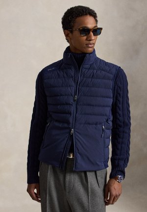 Mann trägt dunkelblaue gesteppte Weste über einem marineblauen Strickpullover mit Zopfmuster, graue Hose, Sonnenbrille und Uhr, steht vor neutralem Hintergrund.