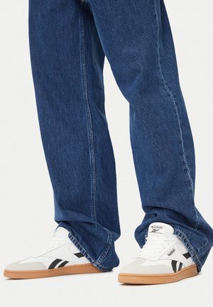 Białe trampki z szarymi i czarnymi akcentami, wyposażone w gumową podeszwę. Noszone z luźnymi ciemnymi dżinsami wykonanymi z teksturowanej tkaniny.