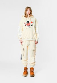 Una persona joven se encuentra de pie vistiendo un hoodie crema con un gráfico rojo, pantalones cargo a juego con cremalleras y botas de cordones color beige, todo contra un fondo blanco.