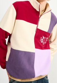 Fleece-pullover med røde, cremefarvede, lilla og lavendel farveblokke; har en høj krave og halv lynlås lukning; logo broderet på fronten.