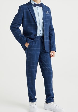 Niño vestido con un traje azul a cuadros con pantalones a juego, camisa azul clara, pajarita azul marino, pañuelo en el bolsillo y zapatillas blancas, de pie con la mano en el bolsillo.