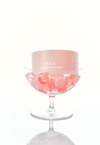 Un récipient cylindrique rose étiqueté « Patch pour les yeux rétinien Abib PDRN » repose dans un bol en verre rempli de cubes de glace roses, reflétant la forme du bol.