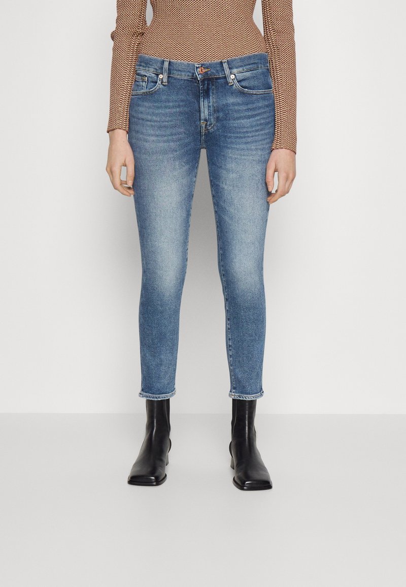 Femme portant un jean slim bleu clair, un haut à manches longues marron à motifs, et des bottines noires, debout devant un fond blanc.