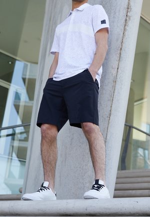 Man in wit gevlekt poloshirt, zwarte korte broek en witte sneakers, staand met handen in de zakken bij een betonnen pilaar buiten een modern gebouw.