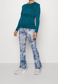 Teal långärmad tröja med gathered midja, parat med blå tie-dye utgående byxor. Modellen bär ljusgrå sneakers.