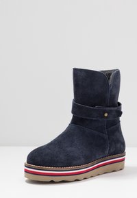 Granatowe zamszowe botki za kostkę z okrągłym noskiem, regulowanym paskiem i żłobioną gumową podeszwą z czerwono-biały paskiem. Miękka podszewka wewnętrzna.
