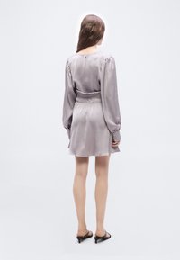 Γυναίκα που στέκεται με την πλάτη προς τον θεατή, φορώντας ένα μακρυμάνικο φόρεμα ανοιχτού μωβ χρώματος και μαύρα ανοιχτά τακούνια kitten, πάνω σε λευκό απλό φόντο.