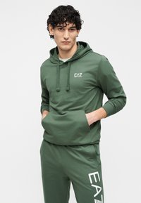 Zelená mikina s kapucňou, predným vreckom, šnúrkami a logom EA7 na hrudi. Dopĺňa ju súprava teplákov so zhodnou potlačou bieleho loga.