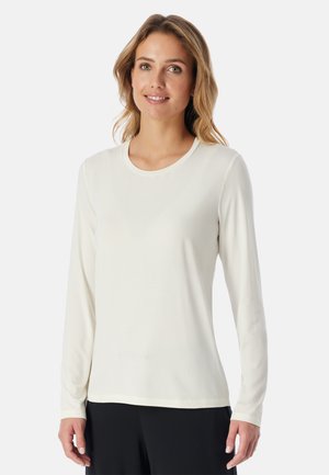 Vrouw draagt een simpel wit shirt met lange mouwen en een zwarte broek, glimlacht met een ontspannen houding tegen een effen lichte achtergrond.