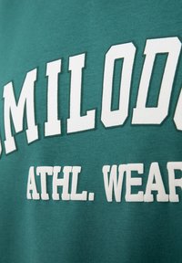 Turkusowy sweatshirt z wyraźnym białym napisem "HAMILOD" oraz "ATHL. WEAR." Miękki materiał o gładkiej fakturze i luźnym kroju.