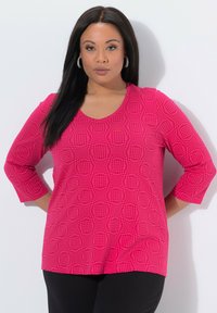 Fuchsia-pinkfarbener Oberteil mit einem strukturierten Kreis-Muster, dreiviertellangen Ärmeln und rundem Ausschnitt, kombiniert mit schwarzer Hose.