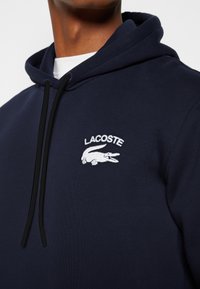Homme portant un sweat à capuche Lacoste bleu marine avec logo blanc et cordons noirs, chemise blanche visible en dessous, gros plan sur la poitrine et le cou.
