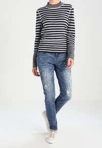 Marineblauer und grauer gestreifter Pullover mit hohem Kragen, kombiniert mit blauen, ausgewaschenen Jeans mit Perlenverzierungen und weißen Turnschuhen.
