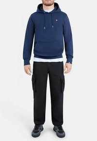 Marineblauer Kapuzenpullover mit Fronttasche; darunter ein weißes Hemd in Lagenoptik. Schwarze Cargo-Hose mit Seitentaschen und dunkle Schuhe.