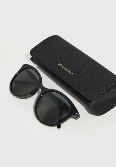 Μαύρα υπερμεγέθη γυαλιά ηλίου με σκοτεινούς φακούς δίπλα σε μια μαύρη θήκη Dolce & Gabbana σε λευκό φόντο.