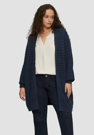 Marineblauwe gebreide cardigan met geribbelde textuur, open voorkant en wijde mouwen, gecombineerd met een witte shirt en donkere jeans.