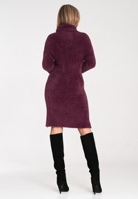 Plüschiges, tiefpurpurnes Strickkleid mit hochgeschlossenem Kragen und langen Ärmeln, das bis zum Knie schmaler wird. Kombiniert mit langen schwarzen Wildlederstiefeln.