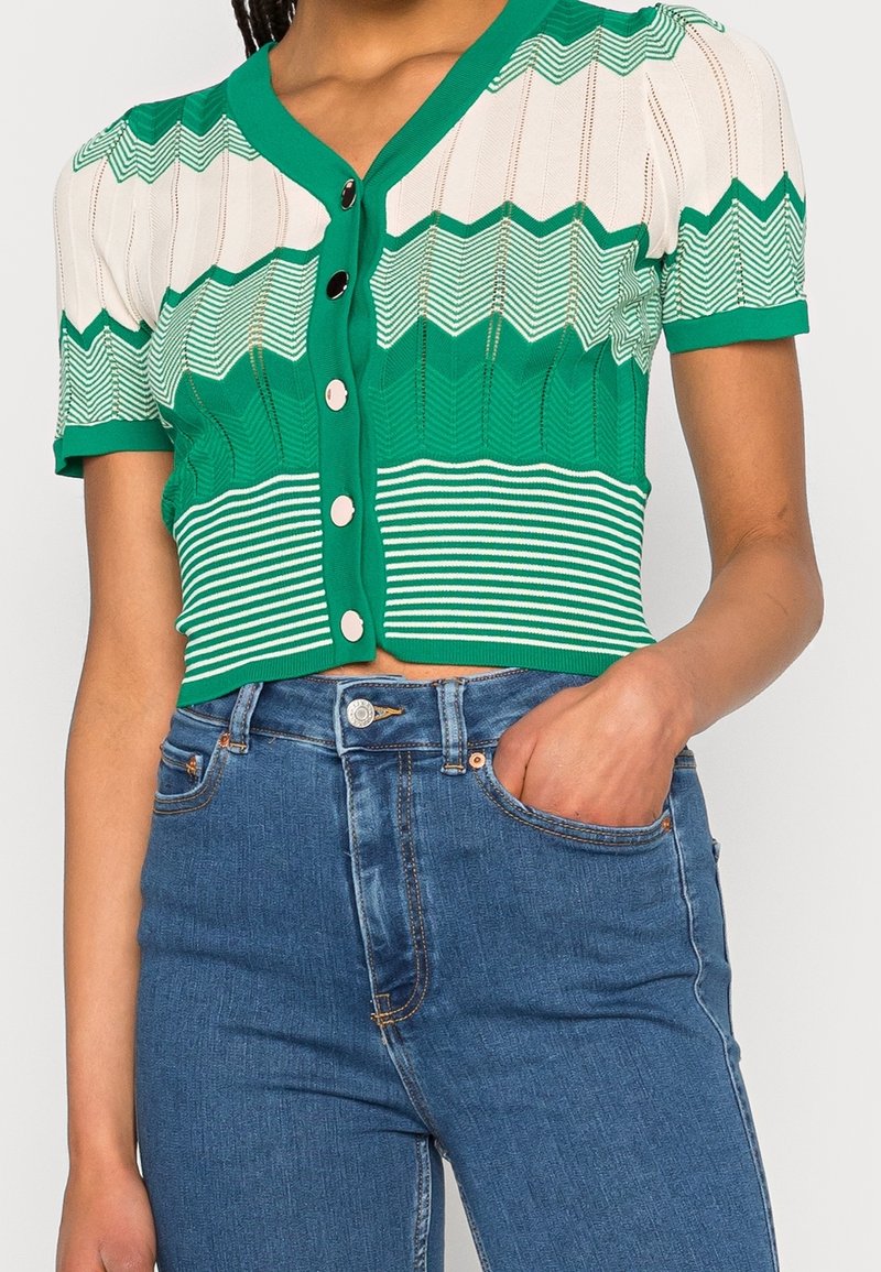 Femme portant un cardigan tricoté vert et blanc à manches courtes avec des boutons, associé à un jean bleu taille haute, main dans la poche avant.