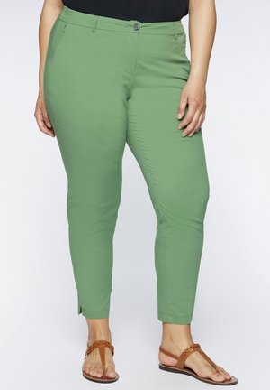 Pantalons slim fit vert clair avec une texture lisse, dotés d'une fermeture par bouton à l'avant et de poches latérales, associés à des sandales marron.