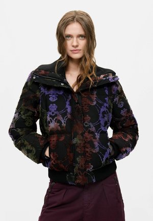 Donna con capelli mossi che indossa una giacca imbottita nera con motivi floreali viola, marroni e verdi, mani nelle tasche e pantaloni viola scuro.