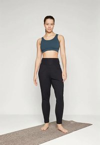 Teal sports bra and black high-waisted leggings, form-fitting, made of stretchable fabric, worn by a standing figure on a yoga mat.