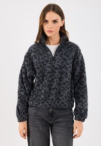 Pulover pufos negru cu un model leopard, guler cu fermoar pe jumătate și mâneci lungi, purtat cu blugi negri. Textură moale.