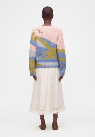 Personne portant un pull à motif abstrait multicolore, une jupe midi beige clair et des chaussures marron à lacets, debout, de dos.
