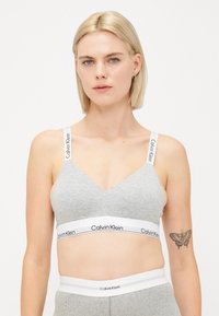 Gray bralette with a V-neck design, featuring a white elastic band with "Calvin Klein" branding. Adjustable shoulder straps and soft texture.