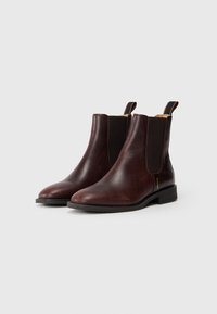 Une paire de bottes Chelsea en cuir marron foncé avec des panneaux latéraux élastiques noirs et des tirettes, sur un fond gris clair uni.