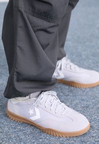 Ljusgrå mockaskor för träning med vit stjärnlogotyp och gummisula, matchade med mörka byxor, på en blå yta.