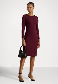 Tumši sarkana pieguļošā kleita ar garām piedurknēm un pogām uz pleciem, komplektā ar melnu rokassomu un melniem sandales stilā augstiem papēžiem uz gaiša fona.