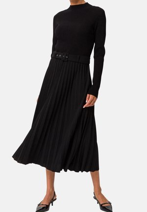 Femme portant un haut noir côtelé à manches longues et une jupe midi plissée avec une ceinture assortie et des chaussures à talons bas noires à bride arrière.
