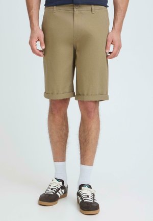 Man draagt beige opgerolde korte broek, witte sokken en zwart-witte Adidas sneakers, staand op een effen achtergrond.