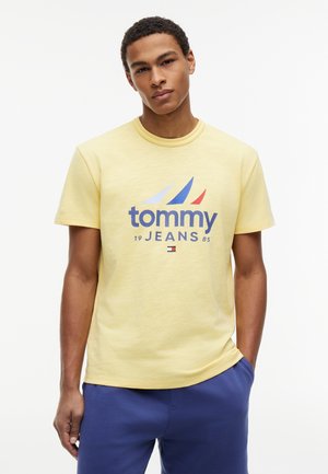 Młody mężczyzna w żółtej koszulce Tommy Jeans z logo i niebieskich spodniach, stojący na jednolitym białym tle.