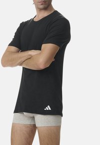 Schwarzes Rundhals-T-Shirt aus Baumwolle mit kurzen Ärmeln und einem weißen Adidas-Logo nahe dem Saum. Das Modell trägt graue Boxershorts.