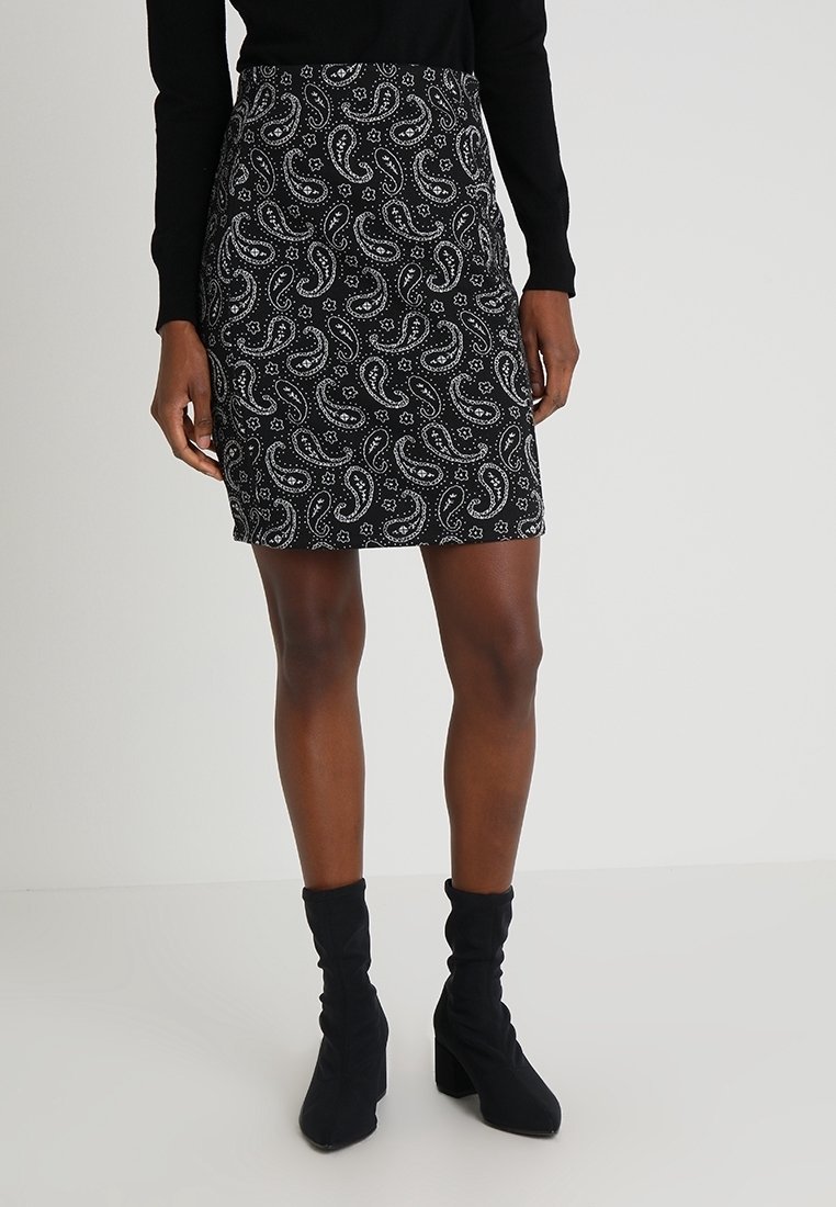 Femme portant une jupe noire avec un motif paisley blanc, un haut noir à manches longues et des bottines noires à talons, debout devant un fond uni.
