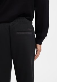 Gros plan d'une personne portant un pantalon noir avec une taille élastique et une poche arrière, main visible reposant à côté.