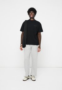 Sort T-shirt med en diskret logo, grå sweatpants og en sort crossbody taske. Hvide og sorte sneakers med tre striber. Ensfarvet baggrund.