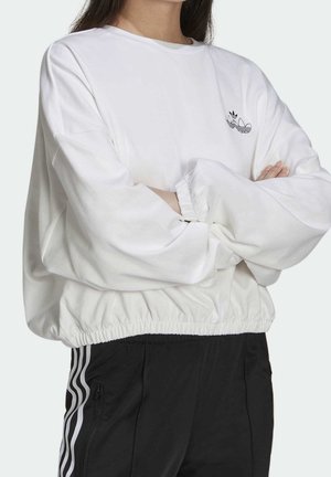 Persoon draagt een wit Adidas-sweatshirt met lange mouwen en logo, en zwarte Adidas-broek met witte strepen, armen gekruist.