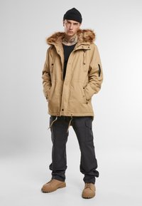 Kaki parka-jakke med hette som har fuskepelsfôr, frontlommer og justerbar snøring. Kombinert med svarte cargo-bukser og beige støvler.