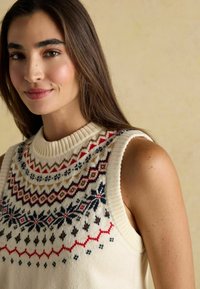 Adītā bez piedurknēm džemperis krēmkrāsā ar rakstainu apkakli, kurā ir sarkani, zili un brūni motīvi. Ribota apkakle un gluda tekstūra.