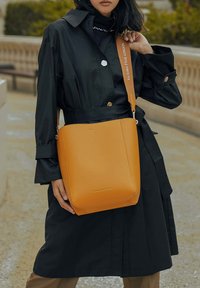 Bruine leren schoudertas met een gladde textuur en minimalistisch ontwerp. Voorzien van een lange strap met ingekerfd merknaam, gedragen door een persoon.