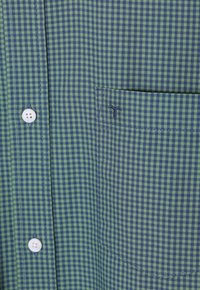 Camisa a cuadros verde y azul marino con cuello abotonado, botones blancos y un bolsillo en el pecho con detalle bordado.