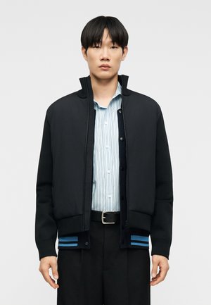 Mladý muž v černé bundě na zip přes světle modrou pruhovanou košili na knoflíky a černé kalhoty s černým opaskem, stojící před jednobarevným pozadím.